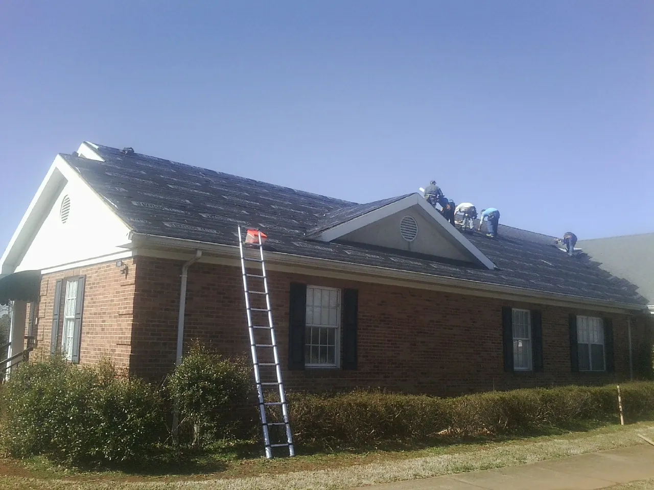 Crew tearing shingles off a brick ranch roof with a ladder set — Emergency Roof Repair work by our crew in Camp Lejeune
