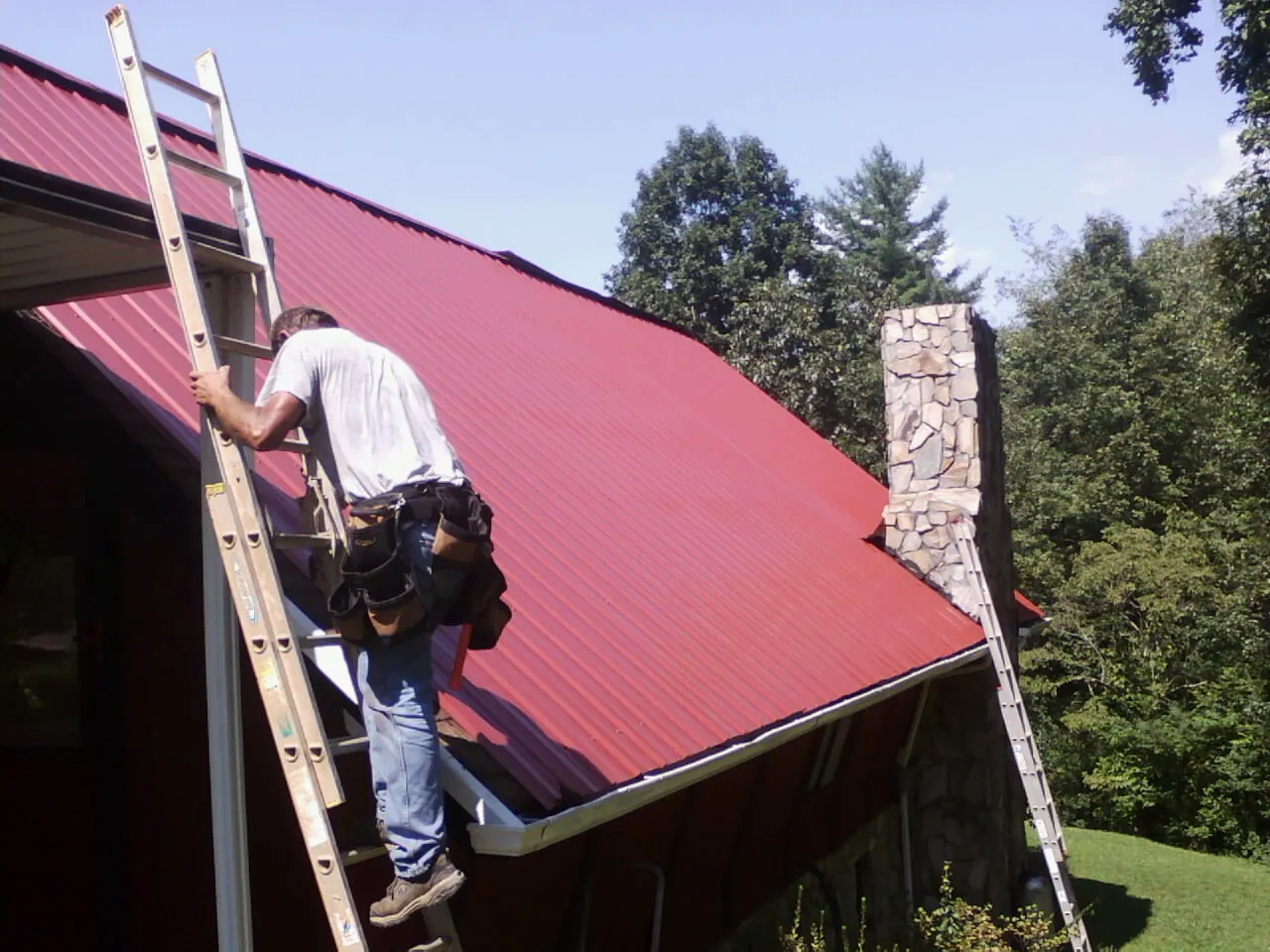 Roofer on a ladder beside a red metal roof and stone chimney — Soffit & Fascia Repair by Roofing Brothers in Camp Lejeune